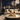 A luxurious modern apartment interior at night with a panoramic window view of a city skyline adorned with exploding fireworks. In the foreground, a round marble coffee table holds a bottle of champagne in a gold ice bucket, a champagne flute, and a vase decorated with fairy lights. A plush white sofa with decorative pillows is visible in the midground, adding to the upscale and festive ambiance.