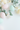 A top-down flat lay shot showcases five pastel-colored Easter eggs scattered across a bright white background, interspersed with fresh eucalyptus branches and tiny white flowers. The eggs are primarily pale pink, mint green, and light yellow, creating a soft, inviting color palette. The composition leaves substantial white space in the lower half of the vertical image, allowing for easy text or graphic overlays.