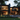 A large, multi-story modern house with dark framing and extensive floor-to-ceiling glass walls is beautifully illuminated from within, set against a backdrop of a dense forest at dusk. The warm glow of the interior lights highlights contemporary furnishings, while lush green foliage and subtle ground lighting frame the exterior, emphasizing its integration with nature and luxurious living.