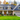 A beautiful, modern two-story family home with a dark grey roof and mixed blue-grey and beige siding, surrounded by vibrant autumn trees displaying brilliant yellow and orange foliage. The house features a welcoming front porch with white columns, a wooden front door, and an attached two-car garage, all set against a lush green lawn and clear, bright sky, conveying a sense of warmth and inviting curb appeal.