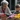 A grandmother with gray hair and red-rimmed glasses, wearing a light pink and white knit cardigan and green gardening gloves, plants flowers with a young boy. The boy, with brown hair, wears a striped hoodie. They are focused on arranging vibrant red, pink, and yellow potted chrysanthemums on a wooden surface, surrounded by loose soil. The scene is outdoors, set against a blurred background of a building and wooden wall, conveying a tender moment of intergenerational learning and connection.