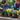 A close-up shot of several potted pansy flowers in various bright colors including yellow, deep purple, and orange, arranged on a rustic wooden table with small piles of dark potting soil. In the softly blurred background, a young boy in a denim shirt and an adult woman in a purple cardigan wearing bright blue gardening gloves are seen actively planting, suggesting a shared, joyful gardening experience. The scene conveys a sense of nurturing, growth, and intergenerational bonding outdoors.