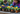 A close-up shot of several potted pansy flowers in various bright colors including yellow, deep purple, and orange, arranged on a rustic wooden table with small piles of dark potting soil. In the softly blurred background, a young boy in a denim shirt and an adult woman in a purple cardigan wearing bright blue gardening gloves are seen actively planting, suggesting a shared, joyful gardening experience. The scene conveys a sense of nurturing, growth, and intergenerational bonding outdoors.