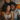 An overhead shot shows a joyful young Black couple lying down, surrounded by cardboard moving boxes and decorative throw pillows. The woman, with curly dark hair and a light patterned top, smiles widely with her eyes closed, nestled beside the man, who also has curly hair and a beard and wears a grey t-shirt, sharing the same expression of deep happiness. They appear content and relaxed in what seems to be their new, yet-to-be-unpacked home, bathed in soft, warm light.