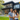 A happy family, including a father, mother, and their toddler, stands smiling on a green lawn in front of a large, dark modern house. The father holds the child, and the mother stands beside him, all looking directly at the camera. The contemporary home features sleek lines, expansive windows with dark frames, and a black and concrete exterior, with lush trees and foliage in the background under soft lighting.