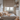 A vertical shot of a modern, cozy child's bedroom decorated in neutral tones. A low-profile bed with cream and peach-colored pillows and a textured blanket faces a window looking out onto city buildings. To the right, built-in shelves and drawers hold a small teddy bear and other items, while a larger teddy bear sits on a fluffy white rug in the foreground.