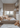 A vertical shot of a modern, cozy child's bedroom decorated in neutral tones. A low-profile bed with cream and peach-colored pillows and a textured blanket faces a window looking out onto city buildings. To the right, built-in shelves and drawers hold a small teddy bear and other items, while a larger teddy bear sits on a fluffy white rug in the foreground.