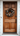 A vertical shot of a warm reddish-brown paneled wooden front door, featuring visible wood grain. A decorative autumnal wreath, made of deep green leaves, white berries, and burnt orange dried flowers, hangs in the upper center. The door is framed by crisp white trim and white horizontal siding, with a black traditional handle and deadbolt on the right, conveying a welcoming and seasonally decorated home entrance.