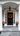 A symmetrical, eye-level shot of the front entrance of an elegant home decorated for autumn. A glossy black paneled door is centered, featuring a vibrant red, orange, and yellow floral wreath, flanked by white-framed sidelights reflecting colorful trees. Two large white fluted columns support a white arched portico, while the surrounding walls are textured grey stone, and the dark grey flagstone porch holds two black urns with matching fall foliage.