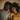 An overhead view shows a young Black couple, a man and a woman, lying next to each other on a textured light brown fabric, looking upwards and smiling. The man wears a gray t-shirt with a patterned pocket and has a beard; the woman, with long curly hair, wears a mustard yellow t-shirt. Their close proximity and gentle smiles suggest warmth and connection.