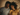 An overhead view shows a young Black couple, a man and a woman, lying next to each other on a textured light brown fabric, looking upwards and smiling. The man wears a gray t-shirt with a patterned pocket and has a beard; the woman, with long curly hair, wears a mustard yellow t-shirt. Their close proximity and gentle smiles suggest warmth and connection.