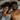 An overhead shot of a young Black couple lying side-by-side on a light beige bed. The woman on the left wears a white tank top and denim vest, smiling with dimples. The man on the right wears a mustard yellow t-shirt and has a beard, also smiling warmly. Both have dark curly hair spread on the pillows, conveying a relaxed and happy mood.