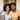 A medium close-up portrait shows a young Black couple smiling brightly at the viewer. The woman, with curly dark hair, wears a light grey t-shirt, while the man, with a beard, wears a mustard yellow t-shirt and has his arm around her. They are seated indoors on a grey couch, in a warmly lit setting with a blurred neutral background.