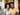 A medium close-up portrait shows a young Black couple smiling brightly at the viewer. The woman, with curly dark hair, wears a light grey t-shirt, while the man, with a beard, wears a mustard yellow t-shirt and has his arm around her. They are seated indoors on a grey couch, in a warmly lit setting with a blurred neutral background.