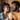A smiling young Black woman with dark curly hair, wearing a light blue denim jacket over a white top, looks towards a young Black man with a beard, who is wearing an orange t-shirt and returning her gaze. Both individuals have warm, authentic smiles on their faces, conveying affection and happiness. The background is softly blurred, showing warm-toned wooden cabinets and a metallic appliance, suggesting a modern home interior.