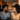 A smiling young Black man and woman look lovingly at each other indoors. The man has a beard and short curly hair, wearing a grey t-shirt with arms crossed. The woman has long curly hair and wears a golden yellow t-shirt. They are in a softly lit kitchen setting with wooden cabinets and a stainless steel refrigerator in the blurred background.