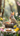 A fluffy, light brown bunny with large ears and a small pink bow sits in a natural nest filled with brightly decorated Easter eggs. The scene is set in a soft-focus, sun-drenched spring garden, with colorful yellow and white flowers, and a blurred dessert on a plate in the foreground. The image evokes a cheerful and whimsical Easter holiday atmosphere.