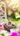 A small, brown and white baby rabbit with a pink bow on its head sits within a natural twig nest. Several pastel-colored Easter eggs are visible around the nest, and various pink, white, and magenta flowers are scattered across a table in the foreground. The background shows a blurred garden scene with a white decorative bench or chair, bright green foliage, and warm, golden sun flare creating a soft bokeh effect.
