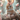 A close-up vertical image shows an adorable light brown and white fluffy baby bunny with a pink polka-dotted bow between its ears, sitting in a rustic bird's nest. Several white eggs with pink and red speckles and one plain light brown egg are visible in the nest next to the bunny. The background and foreground are softly blurred with delicate pink cherry blossoms and branches, bathed in warm, soft light.