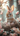 A close-up vertical image shows an adorable light brown and white fluffy baby bunny with a pink polka-dotted bow between its ears, sitting in a rustic bird's nest. Several white eggs with pink and red speckles and one plain light brown egg are visible in the nest next to the bunny. The background and foreground are softly blurred with delicate pink cherry blossoms and branches, bathed in warm, soft light.
