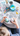 An overhead shot shows a child's hands in a blue knitted sweater holding two decorated Easter eggs. The egg in the left hand is light blue with yellow and pink flowers, while the egg in the right hand has pastel pink and blue swirls with white floral accents. The table around them is cluttered with various pastel eggs, a white paint palette with vibrant colors, paintbrushes in a white vase, and other crafting supplies, all illuminated by soft, bright light.