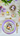 An overhead view displays a beautifully set Easter dining table with a focus on a single place setting. A lavender scalloped charger plate holds a white dinner plate decorated with delicate spring flowers and a small bird's nest containing four pastel Easter eggs. Gold cutlery with floral-patterned handles, along with other decorative eggs, a small ceramic sheep, and fresh purple flowers, are arranged on a rustic white wooden tabletop, conveying a bright, festive, and springtime holiday atmosphere.