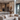 A vertical shot of a luxurious, modern open-concept living room and kitchen. In the foreground is a large beige tufted L-shaped sectional sofa with multiple textured throw pillows, facing a low-profile light taupe coffee table with a dark ribbed vase and faux olive branches. The background reveals a contemporary kitchen with a large white marble-look island, two light brown bar stools, and stainless steel built-in appliances, all illuminated by warm light from modern chandeliers and recessed ceiling lights.