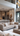 A vertical shot of a luxurious, modern open-concept living room and kitchen. In the foreground is a large beige tufted L-shaped sectional sofa with multiple textured throw pillows, facing a low-profile light taupe coffee table with a dark ribbed vase and faux olive branches. The background reveals a contemporary kitchen with a large white marble-look island, two light brown bar stools, and stainless steel built-in appliances, all illuminated by warm light from modern chandeliers and recessed ceiling lights.
