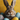 A close-up shot of a fluffy brown and white rabbit with large ears, looking directly at the viewer. The rabbit is nestled in a brown woven basket among several colorful Easter eggs, including speckled blue, speckled white with pink and blue spots, and a solid yellow egg. A vibrant yellow background is softly blurred behind the cheerful scene.