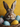 A close-up shot of a fluffy brown and white rabbit with large ears, looking directly at the viewer. The rabbit is nestled in a brown woven basket among several colorful Easter eggs, including speckled blue, speckled white with pink and blue spots, and a solid yellow egg. A vibrant yellow background is softly blurred behind the cheerful scene.