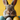 A very close-up, eye-level shot of an adorable brown and grey rabbit's face, with its large dark eyes, pink nose, and long whiskers in sharp focus. Below the rabbit, a blue, a speckled white, and a dotted yellow Easter egg are visible in a brown woven basket, all set against a bright yellow blurred background.