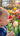 A close-up, side profile of a baby with light hair and fair skin is captured looking intently at a cream-colored tulip in the foreground. The baby's right hand, with small fingers, is slightly extended towards the flower as if to touch it. The background features a vibrant, out-of-focus garden filled with numerous colorful tulips in shades of red, pink, yellow, and purple, all bathed in soft, warm natural light, suggesting a beautiful spring day.