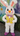 A full-body image of a white Easter bunny mascot standing upright. The bunny has large white ears with pink insides, an orange nose, black eyes, and pink blushed cheeks, wearing a vibrant yellow vest with colorful, dandelion-like patterns, purple buttons, and a purple bow tie featuring small illustrations. It sports bright fuchsia shoes with similar patterns and is positioned outdoors against a background of a large blue flower mural and green foliage, standing on a dark paved surface.