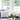 A vibrant vertical image of a modern living room interior. A clear glass vase holds a fresh bouquet of white peonies next to a small golden geometric decoration on a light wooden coffee table. In the background, a dark gray sofa with a white geometric patterned throw pillow and a cream pillow is visible, illuminated by natural light streaming through a large white-framed window looking out onto blurred green trees.