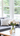A vibrant vertical image of a modern living room interior. A clear glass vase holds a fresh bouquet of white peonies next to a small golden geometric decoration on a light wooden coffee table. In the background, a dark gray sofa with a white geometric patterned throw pillow and a cream pillow is visible, illuminated by natural light streaming through a large white-framed window looking out onto blurred green trees.