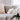 A bouquet of large, white peonies with green leaves is arranged in a clear, fluted glass vase positioned on a white marble coffee table. In the softly blurred background, a beige upholstered sectional sofa with a cream-colored faux fur pillow is visible, bathed in bright, natural light from a nearby window. The scene conveys a clean, elegant, and minimalist interior design aesthetic.