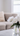 A bouquet of large, white peonies with green leaves is arranged in a clear, fluted glass vase positioned on a white marble coffee table. In the softly blurred background, a beige upholstered sectional sofa with a cream-colored faux fur pillow is visible, bathed in bright, natural light from a nearby window. The scene conveys a clean, elegant, and minimalist interior design aesthetic.
