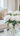 A close-up vertical shot of a beautiful bouquet of white peonies with vibrant green leaves, arranged in a clear glass vase filled with water. The vase sits on a polished, reflective surface, with a softly blurred background showing light-colored furniture and a decorative patterned cushion in a modern, brightly lit room.