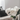 A close-up, vertical shot of a clear glass vase holding several fluffy white peonies with green stems and leaves, placed on a light-colored marble surface. In the blurred background, a light beige sofa with dark grey throw pillows and a draped cream knitted blanket next to an open magazine create a cozy, minimalist home setting illuminated by soft natural light.