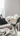 A close-up, vertical shot of a clear glass vase holding several fluffy white peonies with green stems and leaves, placed on a light-colored marble surface. In the blurred background, a light beige sofa with dark grey throw pillows and a draped cream knitted blanket next to an open magazine create a cozy, minimalist home setting illuminated by soft natural light.