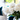 A close-up, vertical shot of a beautiful bouquet of white peonies with green stems, arranged in a clear glass vase on a white table. The background is a brightly lit, out-of-focus modern living room, featuring a cream sofa with a deep teal velvet cushion and a large window looking out onto green foliage. The image evokes a feeling of freshness, elegance, and serene luxury.