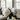 A clear glass vase holds a lush bouquet of fluffy white peonies with vibrant green leaves and visible stems in water, placed on a reflective table. In the softly blurred background, a bright living room features a light-colored sofa on the left and large windows showcasing green trees and horizontal blinds on the right, all illuminated by gentle natural light.