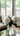 A clear glass vase holds a lush bouquet of fluffy white peonies with vibrant green leaves and visible stems in water, placed on a reflective table. In the softly blurred background, a bright living room features a light-colored sofa on the left and large windows showcasing green trees and horizontal blinds on the right, all illuminated by gentle natural light.