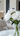 A close-up vertical shot of a beautiful bouquet of white peonies in a clear glass vase, sitting on a white marble table. The large, layered white flower blooms have visible green stems and leaves, with a blurred modern minimalist interior including furniture and light walls in the background. The overall image conveys a sense of clean elegance and fresh sophistication.