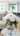 A clear glass vase holding a vibrant bouquet of lush white peonies with green leaves sits on a round white marble-top table with gold trim. In the softly blurred background, a bright, modern open-plan living space is visible, featuring a neutral-colored sofa with a sage green pillow, large windows with white curtains, and a contemporary kitchen area with dark bar stools. The entire scene is bathed in natural light, conveying an atmosphere of freshness and serene elegance.