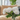 A vibrant bouquet of white peonies with green stems and leaves sits in a clear glass vase on a round, black-rimmed reflective glass coffee table. In the soft-focused background, a beige sofa is visible, along with bright natural light from a window and a large green houseplant on the right, creating a serene and modern home interior scene.
