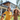 A joyful young woman with dark hair in a yellow t-shirt and two young boys are running and laughing in a sun-drenched green backyard. The older boy, with abundant curly brown hair, wears a brown t-shirt and holds the woman's hand, while the younger boy is partially visible to the left. A stylish modern house with white walls and wood paneling is visible in the blurred background under a clear sky.