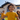 A low-angle shot features three young children with curly hair smiling brightly while playing outdoors. The child in the foreground, wearing a yellow t-shirt, looks directly at the camera with a wide, open-mouthed smile. Behind them, two other children are in soft focus, standing on a green lawn in front of a modern, light-colored house with a dark roof and large windows, under a clear blue sky.
