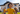 A low-angle shot features three young children with curly hair smiling brightly while playing outdoors. The child in the foreground, wearing a yellow t-shirt, looks directly at the camera with a wide, open-mouthed smile. Behind them, two other children are in soft focus, standing on a green lawn in front of a modern, light-colored house with a dark roof and large windows, under a clear blue sky.