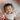 An overhead shot of a cheerful baby with dark hair, lying on white bedding and smiling broadly with an open mouth. The baby is wearing a plain white short-sleeved onesie. On either side of the baby's head, the bedding features sections of white fabric printed with a repeating pattern of bright red strawberries and small green stems, creating a colorful and cozy frame.